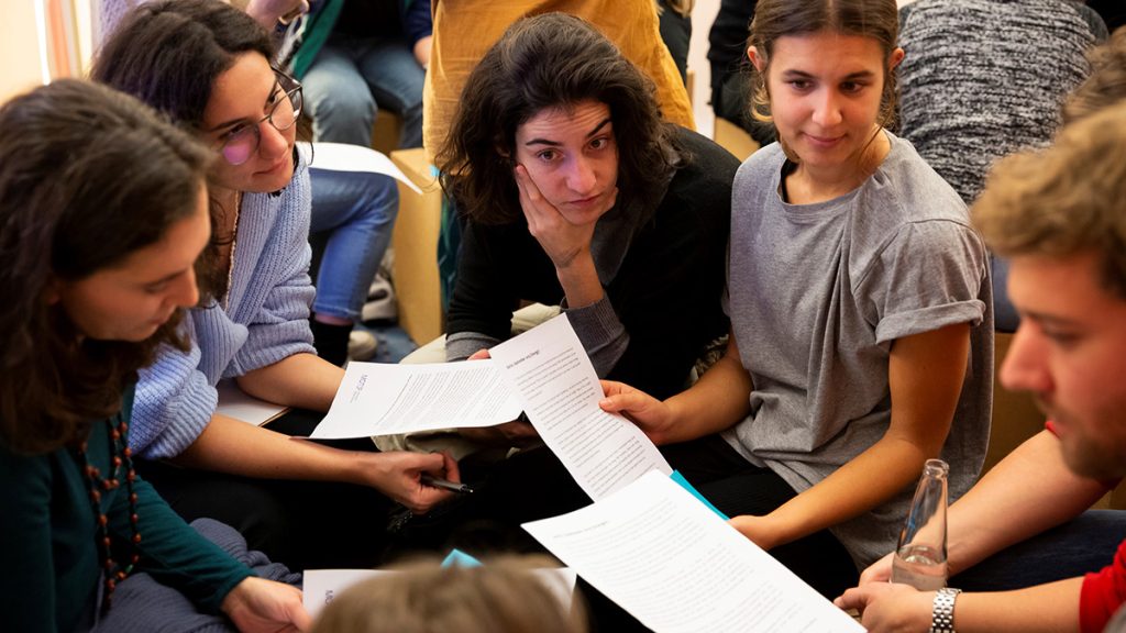Fünf junge, neugierig blickende Personen beugen sich bei einem Workshop gemeinsam über Textdokumente.
