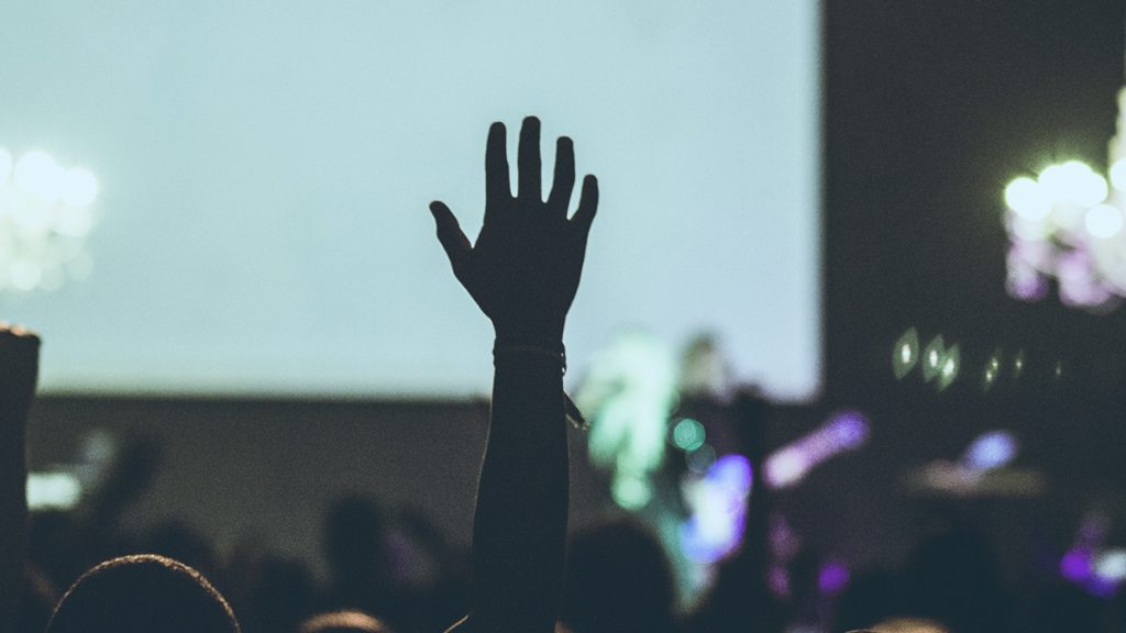 Eine Person hebt eine Hand aus dem dunklen Publikumsbereich eines Konzerts. Im Hintergrund erstrahlt die Bühne mit Band.