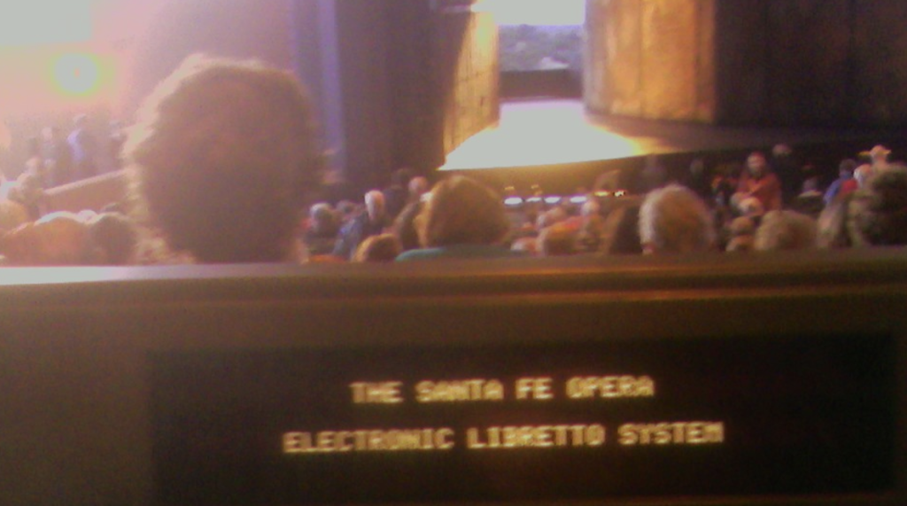 Die Oberkante eines Theaterstuhls der Santa Fe Opera, in den ein Untertitelsystem verbaut ist. Im Hintergrund ein Theaterraum mit Bühne. 