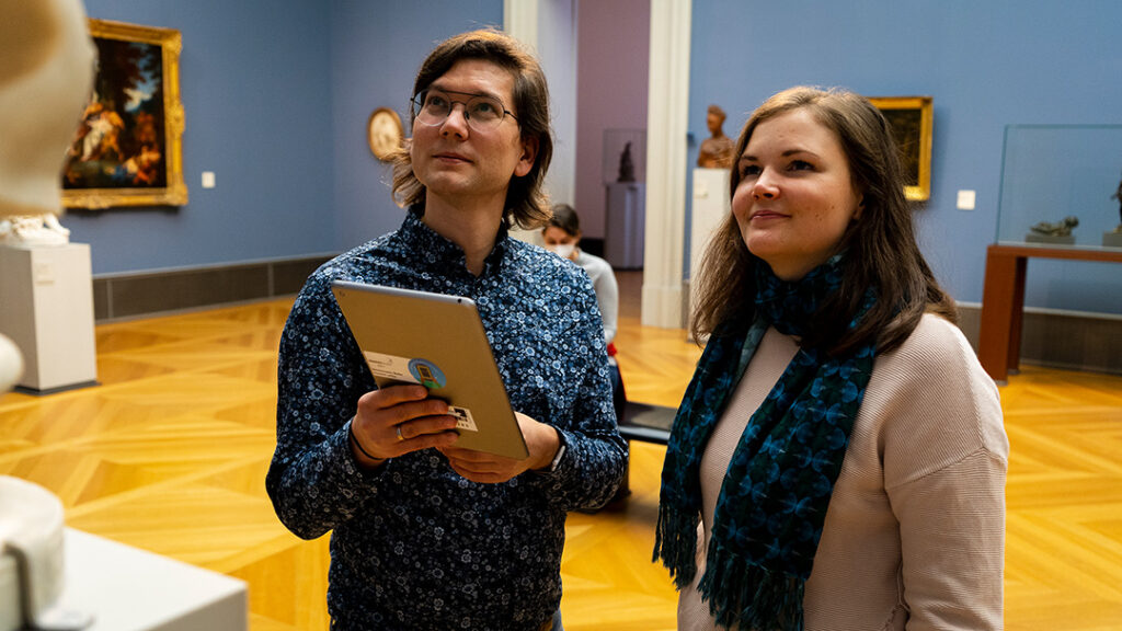 Ein junger Mann und eine junge Frau betrachten in einem Museum eine Statue, die nicht direkt gezeigt wird. Der Mann hält ein iPad.