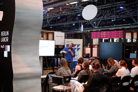 Gezeigt wird eine Vortragssituation im Berlin Labor auf der republica. Das Publikum sitzt mit dem Rücken zur Kamera; der Referent Georg Werner von "ZusammenPlayer" steht in der Mitte des Bildes neben einem Bildschirm und spricht in ein Mikrofon.