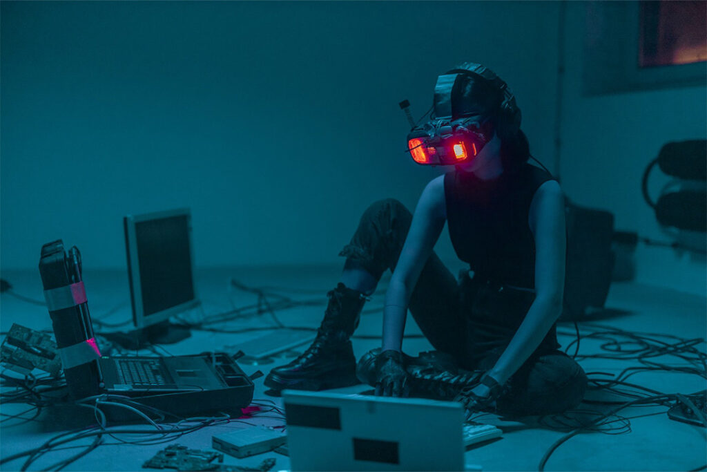 In einem abgedunkelten Zimmer sitzt eine Person mit Virtual Reality-Brille auf dem Kopf auf dem Boden. Vor ihr stehen verschiedene IT-Gerätschaften und sie tippt auf eine Tastatur ein.
