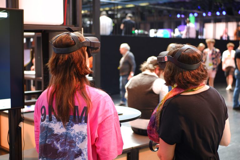 Zwei junge Personen stehen mit dem Rücken zur Kamera in einer Messehalle und tragen VR-Brillen. Im Hintergrund laufen weitere Besucher:innen durch das Bild.