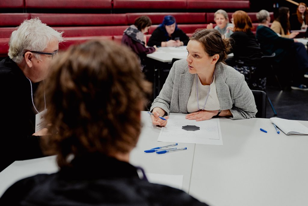 Mehrere Teilnehmende sitzen an einem Tisch und diskutieren. Eine weiblich gelesene Person in grauem Blazer ist konzentriert und spricht mit der Person, die daneben sitzt. Im Hintergrund arbeiten weitere Personen in kleinen Gruppen.