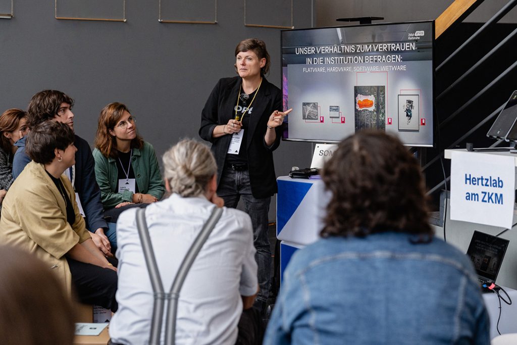 Eine Präsentation in der Galerie, bei der die Sprecherin Tina Lorenz auf einen Bildschirm zeigt, auf dem der Text „Unser Verhältnis zum Vertrauen in die Institution befragen: Flatware, Hardware, Software, Wetware“ steht. Mehrere Personen hören aufmerksam zu und wirken nachdenklich und interessiert.