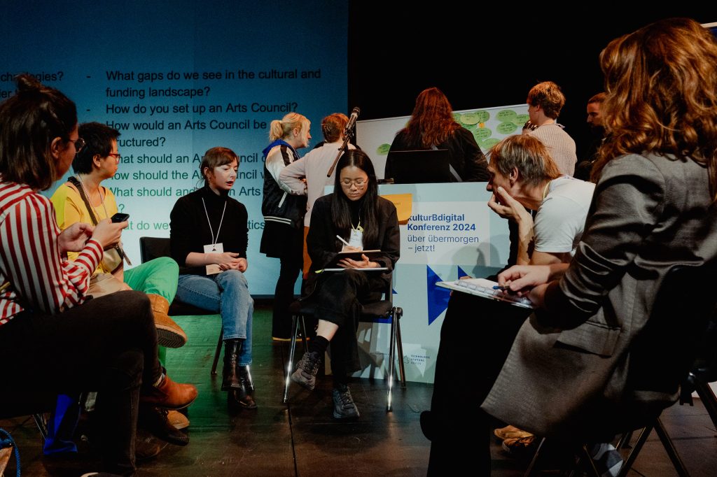 Eine Gruppe sitzt diskutierend im Kreis auf der Bühne der kulturBdigital-Konferenz. Im Hintergrund steht eine weitere Gruppe Konferenz-Teilnehmer:innen um ein Whiteboard herum, auf dem viele grüne Zettel kleben.