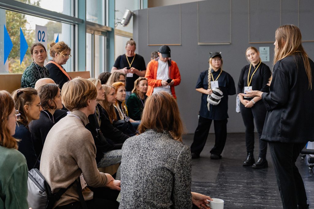 Eine Session in der Galerie, bei der eine Referentin spricht und eine Gruppe von Teilnehmenden aufmerksam zuhört. Einige Personen wirken nachdenklich, andere notieren sich möglicherweise Informationen. Die Umgebung ist offen, mit großen Fenstern im Hintergrund.