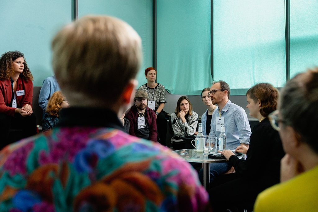 Eine Gruppe von Konferenzteilnehmenden sitzt in einem Raum mit grünen Vorhängen. Andreas Krüger spricht, während die anderen aufmerksam zuhören. Die Atmosphäre ist konzentriert und diskussionsfreudig.