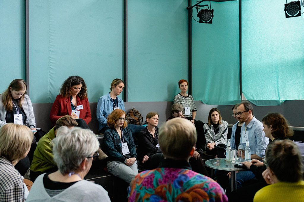 Mehrere Personen sitzen in einer Diskussionsrunde, in einem Raum mit grünen Vorhängen. Einige Teilnehmende machen sich Notizen, während andere zuhören. Die Stimmung ist konzentriert und ernsthaft.