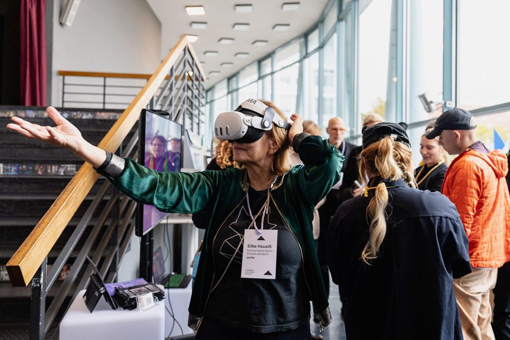 Eine Frau trägt eine VR-Brille im Foyer und hebt die Arme, während andere Teilnehmende im Hintergrund diskutieren. Auf ihrem Namensschild steht „Silke Haueiß“. Die Szene wirkt lebendig und neugierig, während die Person die VR-Erfahrung genießt