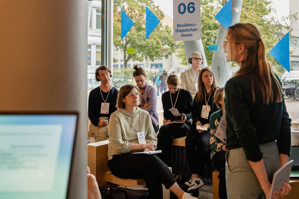 Eine weiblich gelesene Person leitet die Diskussion in der Session. Die Anwesenden, die auf Stühlen und Bänken sitzen, hören aufmerksam zu und machen sich gelegentlich Notizen. Dekorationselemente und Beschriftungen zur Session sind an den Fenstern sichtbar.