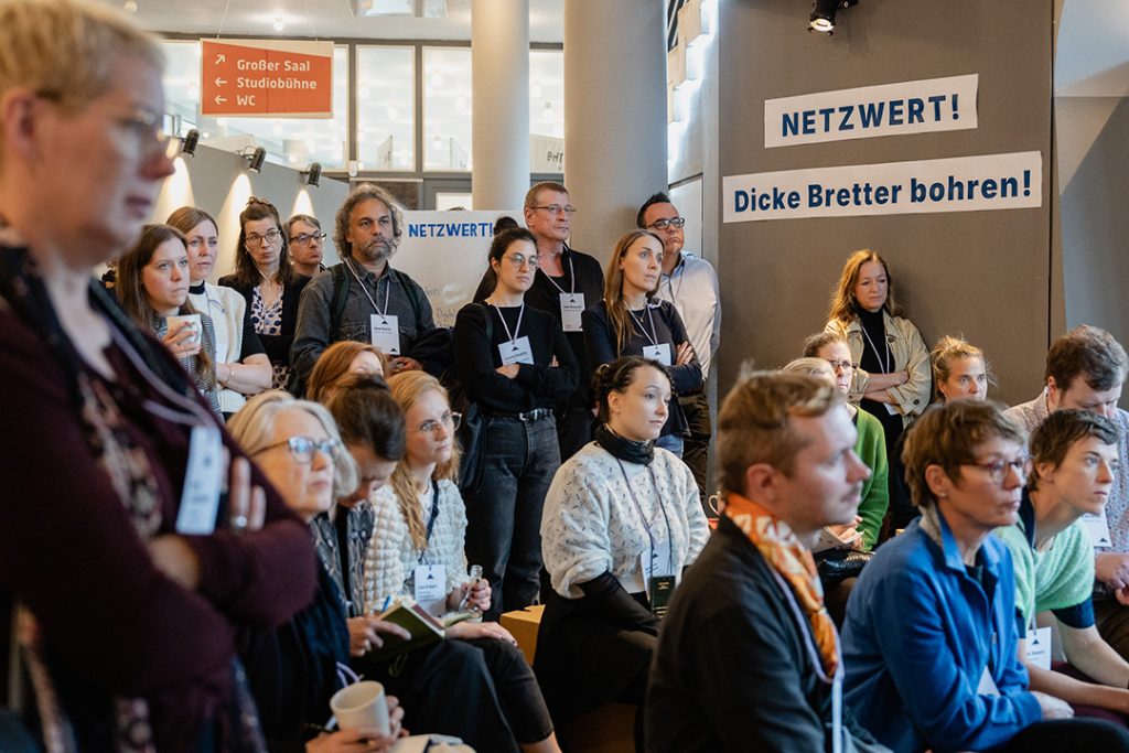 Im Foyer der Konferenz verfolgen Teilnehmende aufmerksam die Session von Resillienz-Dispatcher:innen. Die Teilnehmenden stehen und sitzen dicht gedrängt, schauen ernst und konzentriert. Im Hintergrund ist der Slogan „NETZWERT! Dicke Bretter bohren“ sichtbar.