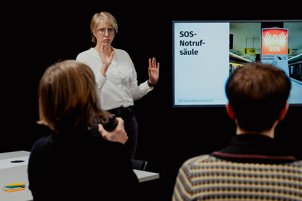 Jolanta Paliszewska gebärdet vor zwei Workshop-Teilnehmenden. Ihr Fallbeispiel ist auf einem Bildschirm zu sehen: Eine SOS-Notrufsäule.