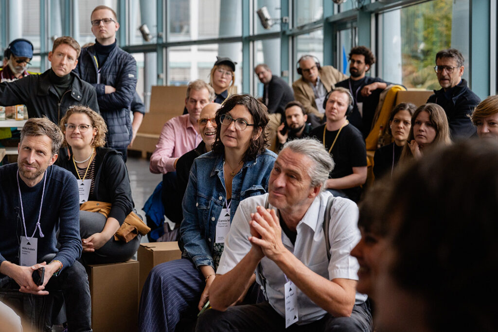 Eine große Gruppe von Konferenzteilnehmenden sitzt im Foyer und lauscht aufmerksam der Session. Einige lächeln, während andere nachdenklich wirken.