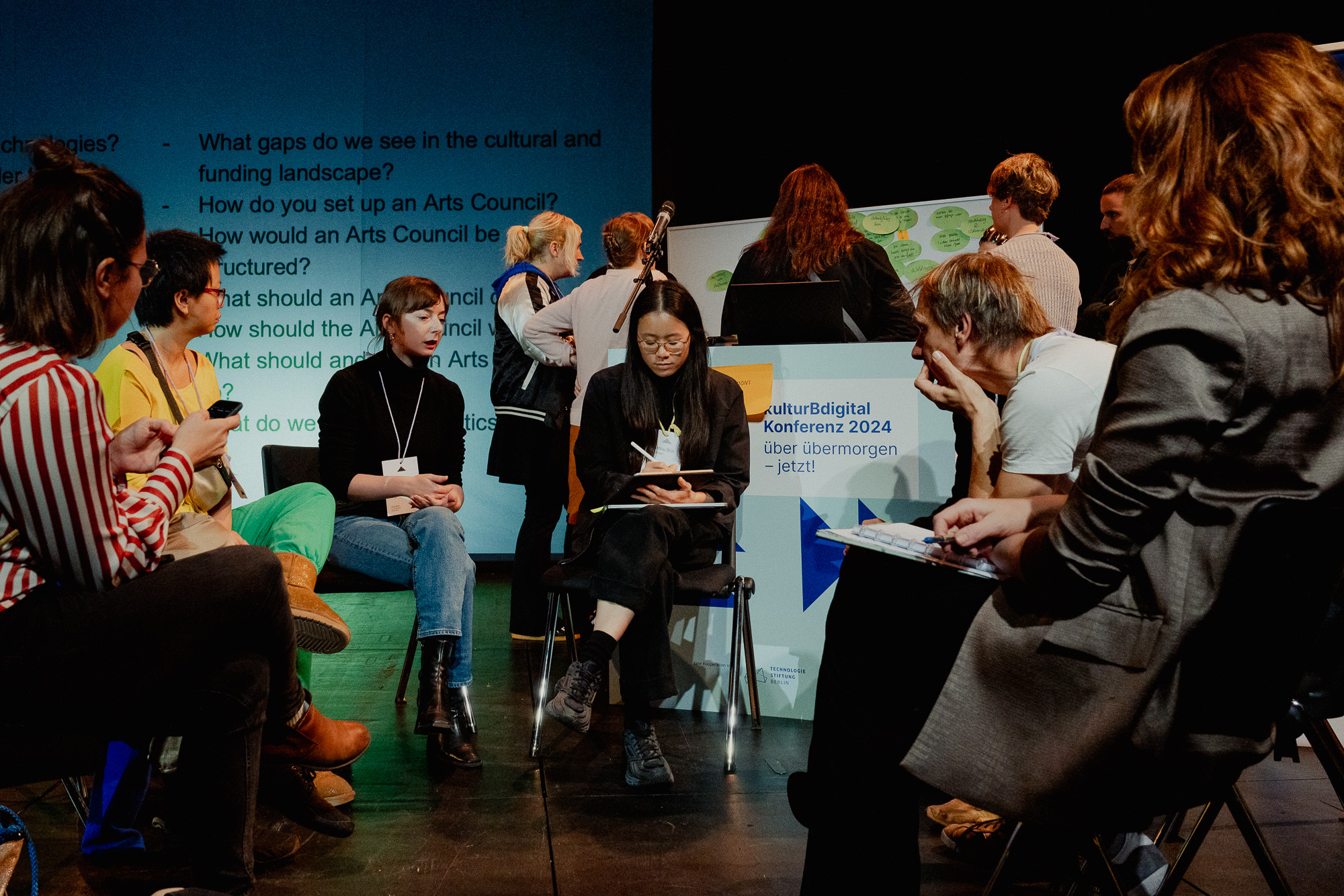 11 Konferenzteilnehmende diskutieren auf der Bühne und machen sich Notizen zu den im Hintergrund projizierten Diskussionsfragen.