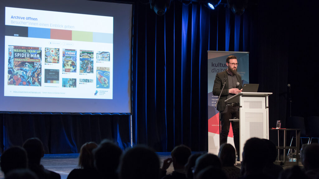 Dr. Sebastian Meier spricht am Rednerpult. Die Präsentation neben ihm fordert Archive öffnen und zeigt Bilder historischer Comicausgaben.