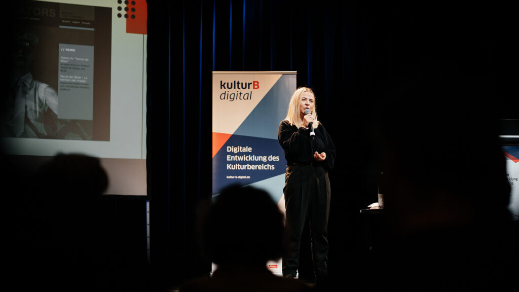 Sabrina Apitz spricht vor einem kulturBdigital-Banner in den dunkel ausgeleuchteten Publikumsraum.