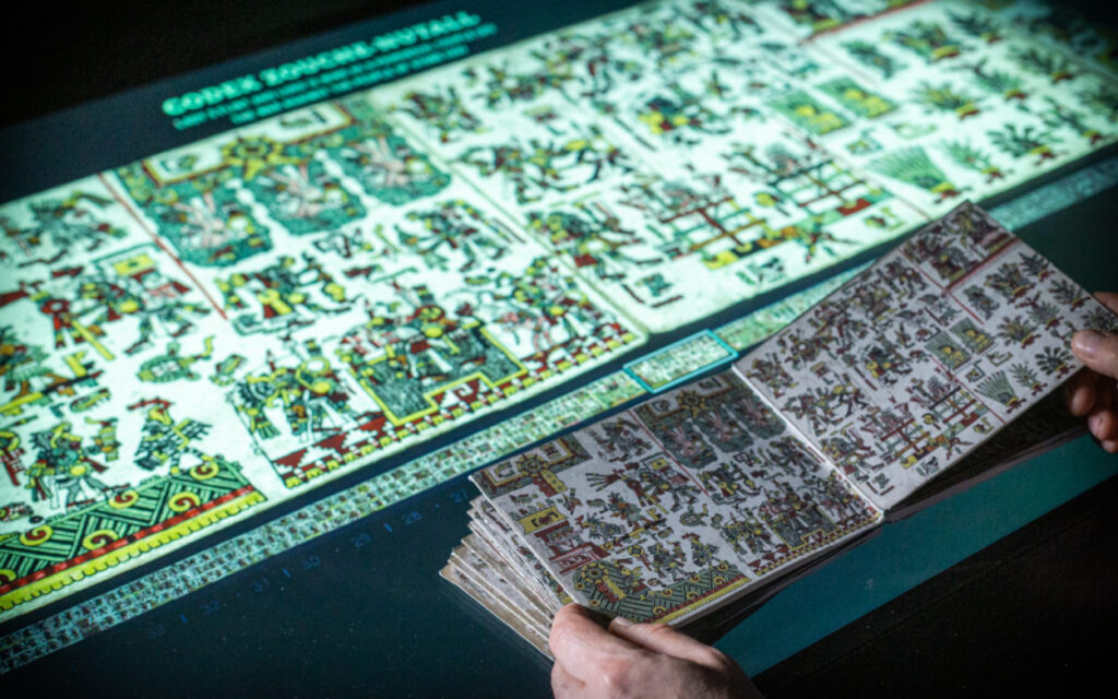 Detailansicht des vergößerten, digitalisierten Schriftstücks mit mehrfarbigen Glyphen, ein Textsegment ist mit Rahmen markiert. Davor liegt eine Replik des kleineren Originalkodex.