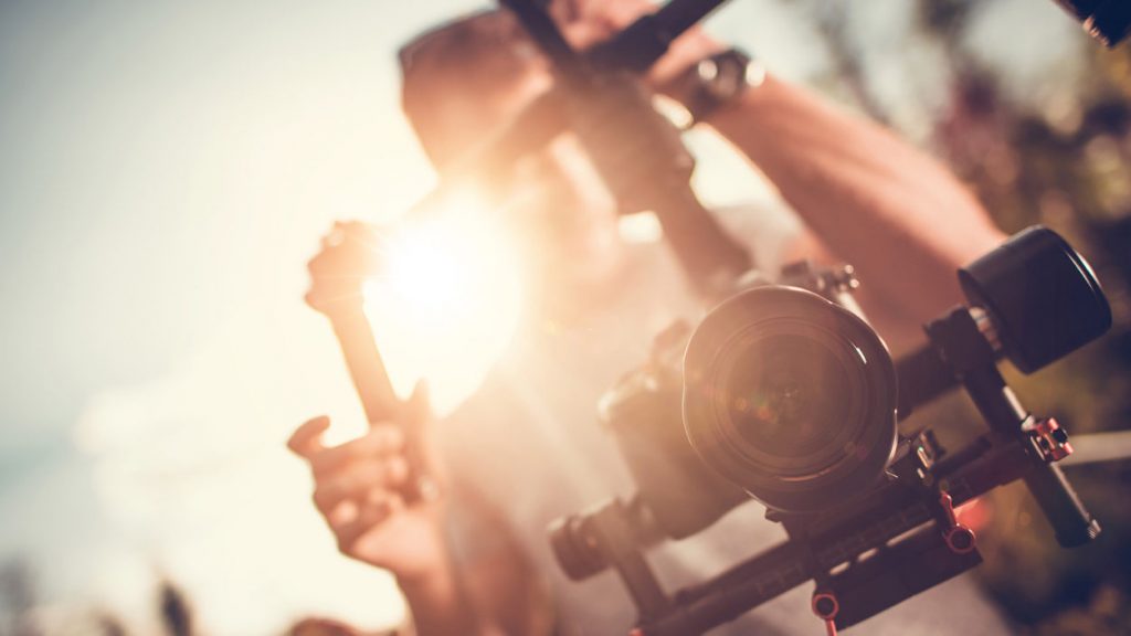 Eine Person hält eine Digitalkamera, die auf ein Gimbal montiert ist. Das Gesicht der Person ist von der Sonne umrahmt.