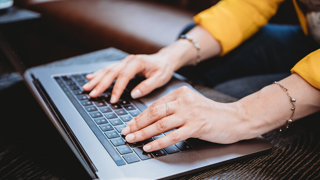 Eine Person mit zierlichen Händen, Ring und Armbändern tippt auf einem Laptop.