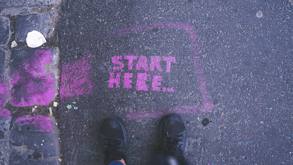 Die Füße einer Person stehen an einem pinken Boden-Graffiti mit dem Text Start Here.