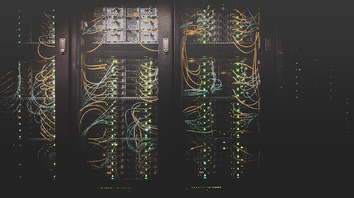 Blick in einen dunklen Serverraum mit grün leuchtenden Server-Lämpchen und wilder Verkabelung.