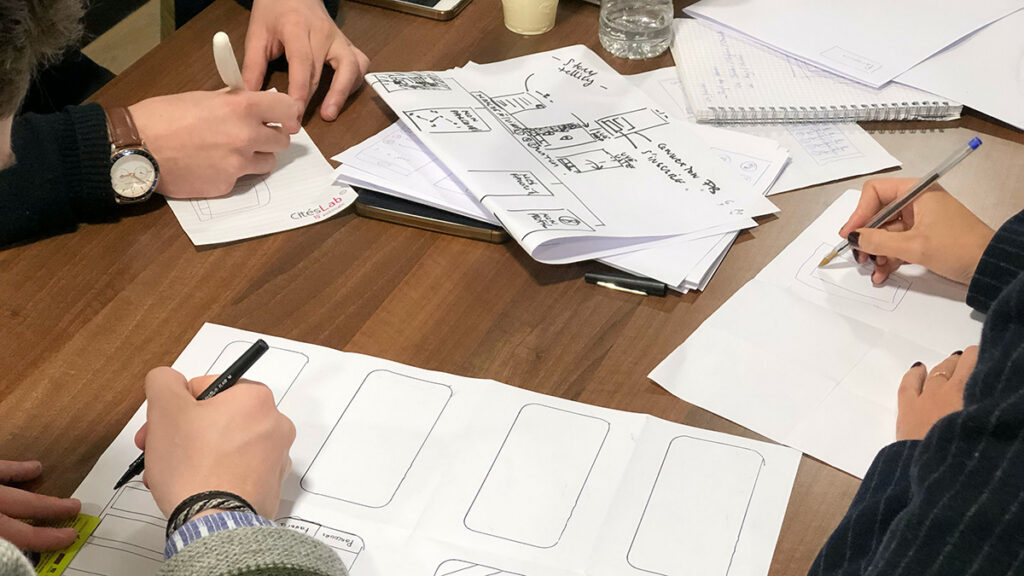 Drei Personen, von denen nur die Hände zu sehen sind, zeichnen Wireframes einer Smartphone-App auf Papierbögen.