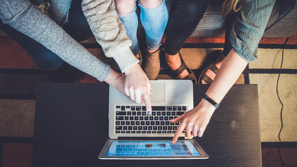 Drei Personen zeigen auf einen Laptop-Bildschirm. Eine Person hat blau lackierte Nägel und eine Jeans mit Knielöchern.
