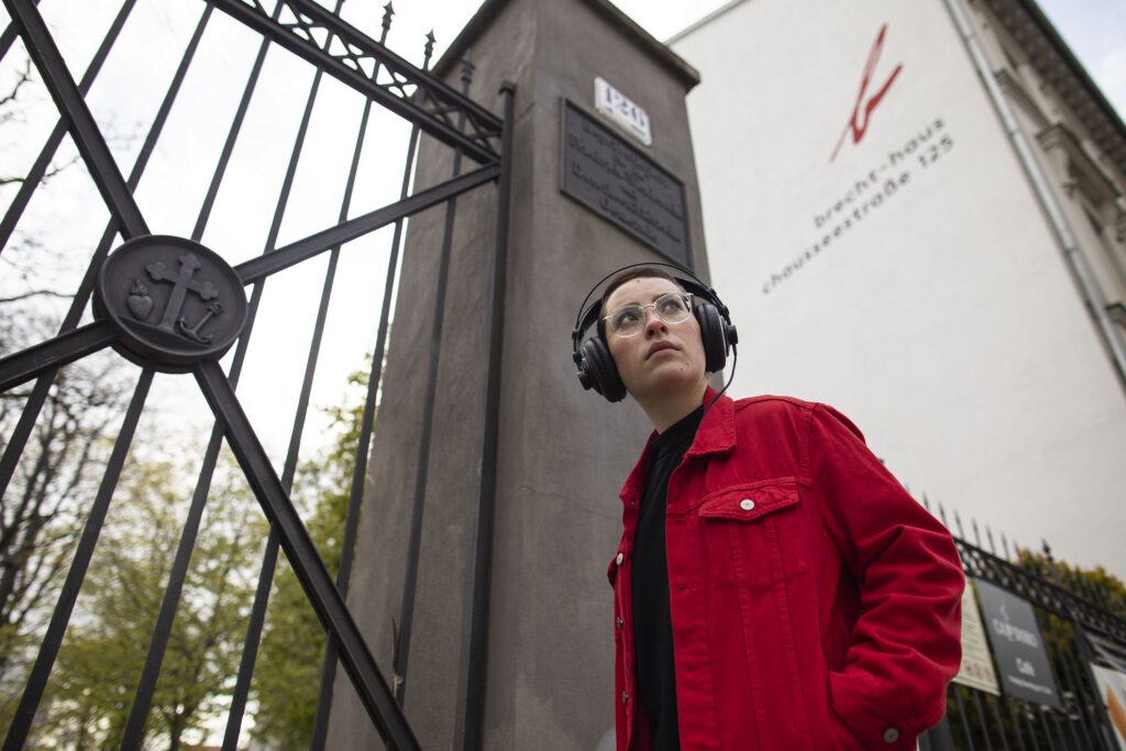 Eine junge Person mit Brille, roter Jacke und aufgesetzten Kopfhörern schaut vor einem Tor in Berlin in den Stadtraum.