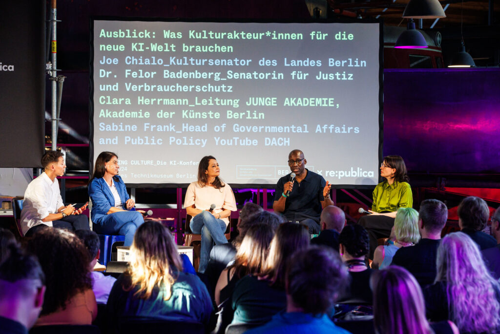 Blick aus der Mitte des Publikums auf die Bühne der KI-Konferenz. Hier sitzen Moderator:in AC Coppens, Sabine Frank (YouTube DACH), Senatorin Felor Badenberg, Senator Joe Chialo und Clara Herrmann (Akademie der Künste). Hinter ihnen eine Präsentation mit den Namen der Diskutant:innen und dem Titel der Podiumsdiskussion: Ausblick. Was Kulturakteur:innen für die neue KI-Welt brauchen.