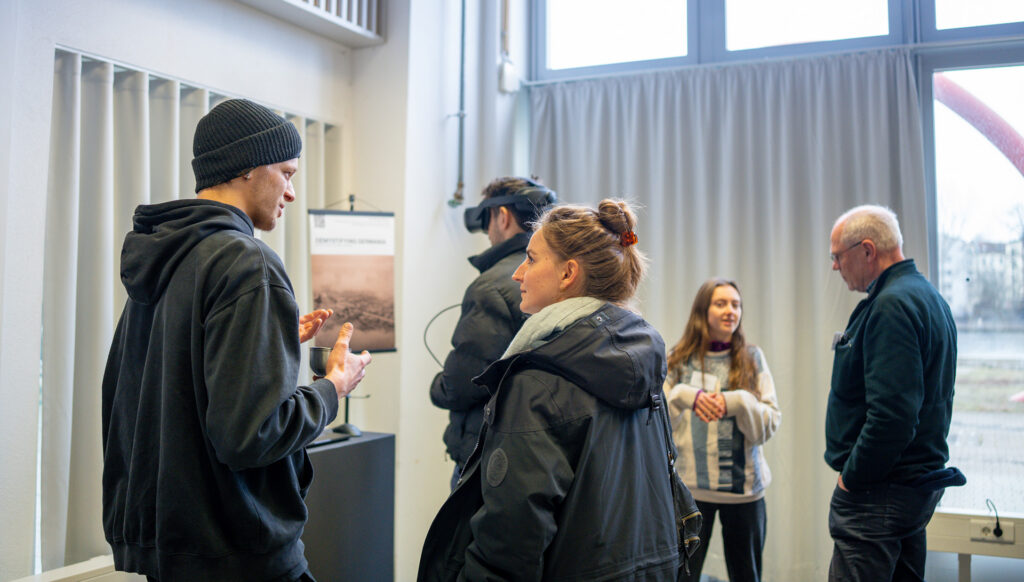 Ein junger Mann steht vor der Präsentation seines XR-Projekts und spricht mit einer jungen Frau. Im Hintergrund stehen weitere Personen in einer kleinen Gruppe zusammen, ein Mann trägt eine VR-Brille.