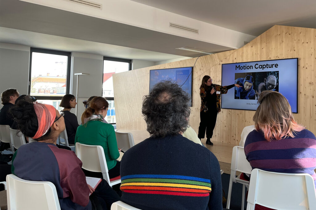 Xenia Kitaeva im Veranstaltungsraum der Technologiestiftung, im Vordergrund das Publikum. Sie zeigt auf eine Präsentation, die mit Motion Capture betitelt ist und links Schauspieler Andy Serkis sowie rechts den von ihm gespielten Gollum aus dem Film Herr der Ringe-Filmen zeigt.