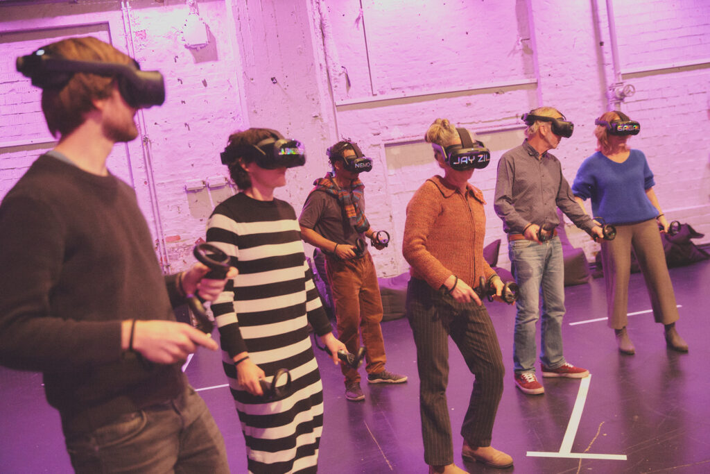 Foto aus der interaktiven VR-Produktion Breaking Virtual. Teilnehmer:innen stehen in einem violett ausgeleuchteten Raum. Sie tragen VR-Headsets und halten Controller in den Händen.