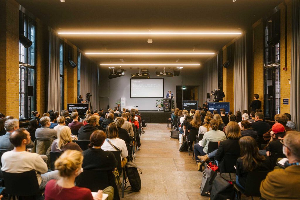 Blick in den Vortragssaal während der kulturBdigital-Konferenz 2022 im bUm in Berlin: Das Publikum blickt zur Bühne, wo Paul Spies, Direktor des Stadtmuseums Berlin gerade an einem Pult spricht.