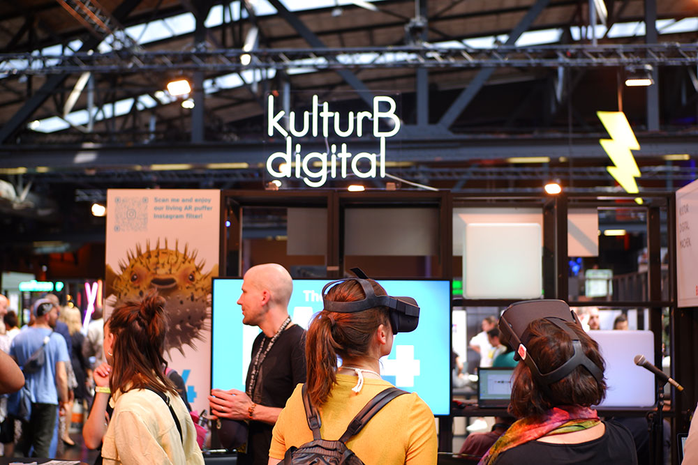 Publikum am kulturBdigital-Stand auf der republica 2023: Zwei Frauen im Vordergrund tragen VR-Brillen. Hinter ihnen sind weitere Personen im Gespräch. In der oberen Mitte des Fotos leuchtet ein LED-Schild mit dem Schriftzug kulturBdigital.