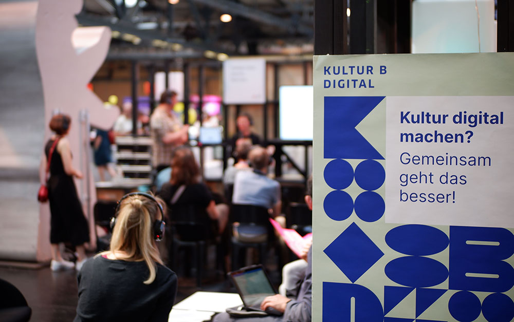 Vorne rechts im Bild ist ein Schild von kulturBdigital mit dem Schriftzug "Kultur digital machen? Gemeinsam geht das besser!" fokussiert. Links im Hintergrund sind unscharf Menschen zu sehen, die einem Vortrag zuhören.