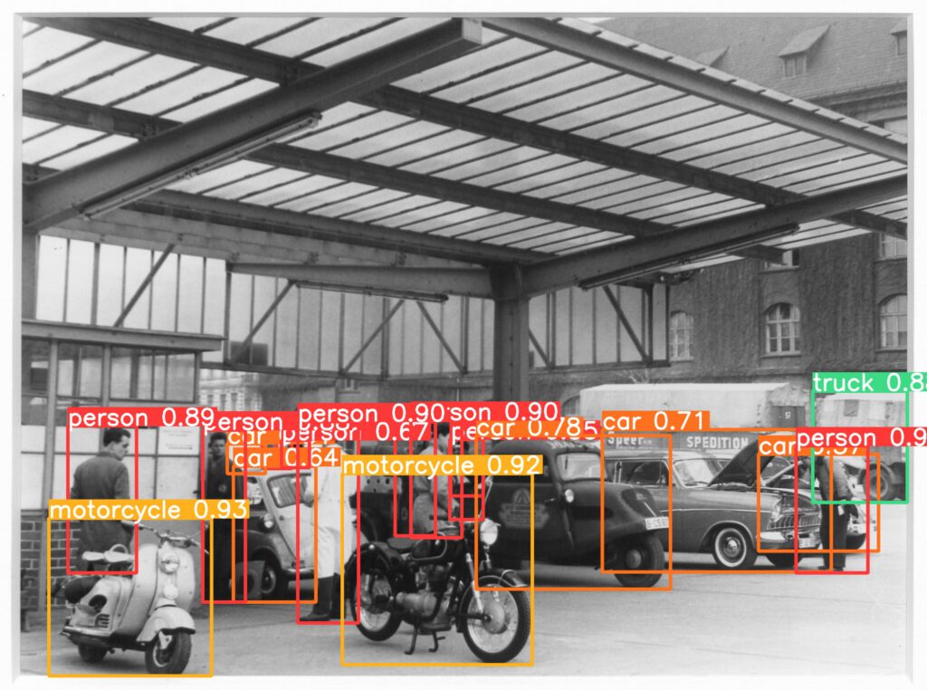 Fotografie der Außenanlage einer Berliner Werkstatt. Es warten Mopeds, Autos. Ein Motorrad und ein Moped werden als "motorcycle", Autos als "car" erkannt, auch um viele Menschen herum befinden sich rechteckige Rahmen mit dem korrekten Schlagwort "person" und hohen confidence-Werten bis 90 Prozent. Ein Lieferwagen wird als "truck" erkannt".
