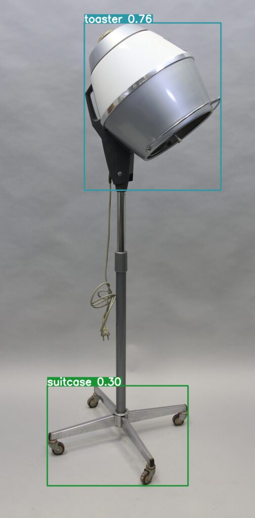 Haartrockner der Firma Wella, Modell "Wella Solar". Ständer auf vier Füßen mit Rollen; höhenverstellbarer Arm; halbkugelförmige Haube aus grau-metallicfarbenem und weißem Blech gefertigt mit glänzenden silberfarbenen Details; rückseitig Kabelanschluss angebracht. Um die Haube herum befindet sich ein rechteckiger Rahmen mit dem Schlagwort "toaster" als Ergebnis der Bilderkennung. Die Wahrscheinlichkeit für den Fund wird mit 76 Prozent angegeben. Um die Füße herum befindet sich ein rechteckiger Rahmen mit dem Schlagwort "suitcase". Die Wahrscheinlichkeit für den Fund: 30 Prozent.