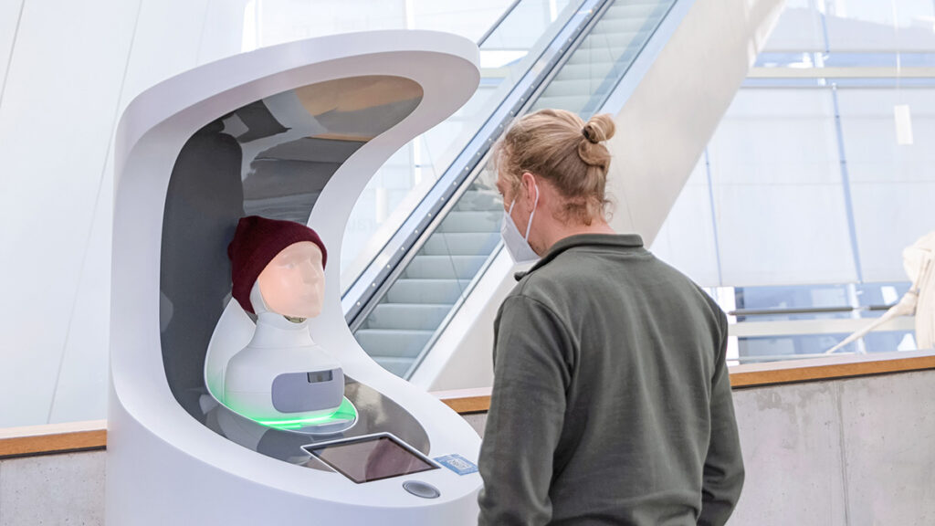 Besucher im OZEANEUM schaut auf einen Service-Roboter mit menschlichen Gesichtszügen und Wollmütze.