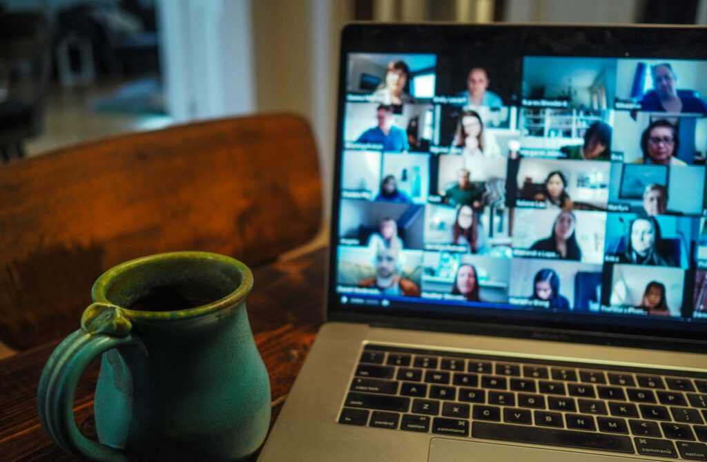 Ein Laptop auf einem Holztisch zeigt eine Videokonferenz mit vielen Teilnehmenden. Im Vordergrund steht eine grün-blaue Keramiktasse.