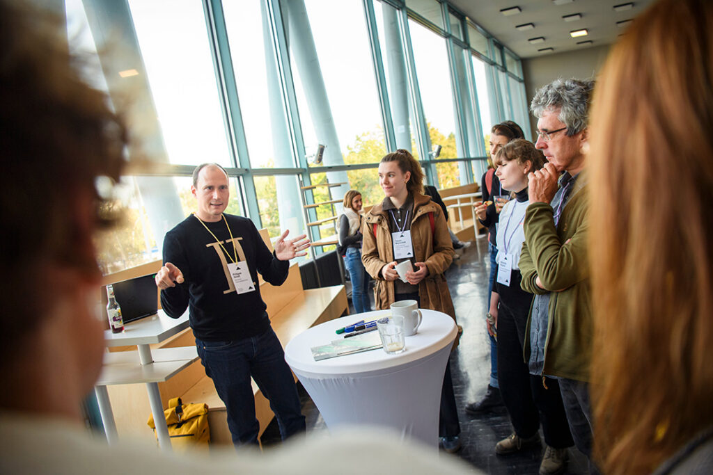Philippe Genêt berichtet vor Gästen der kulturBdigital-Konferenz von Ergebnissen des Coding DaVinci-Projekts. Die Gruppe steht im lichtdurchfluteten Foyer des ATZE-Musiktheater.
