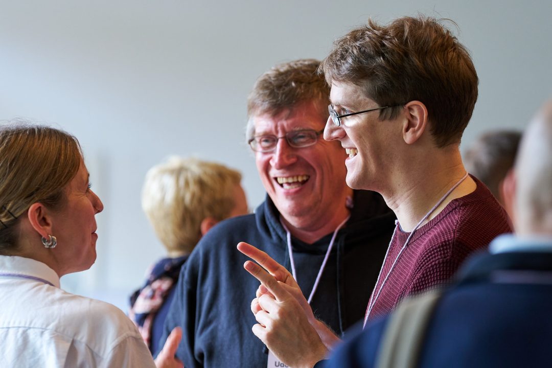 Drei Teilnehmende stehen im Foyer, schauen belustigt und lachen einander an. Eine Person gestikuliert.