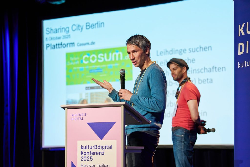 Nikolai Wolfert steht am Rednerpult und spricht ins Mikro. Jan Philip Steimel steht lächelnd hinter ihm. Im Hintergrund steht: Sharing City Berlin.