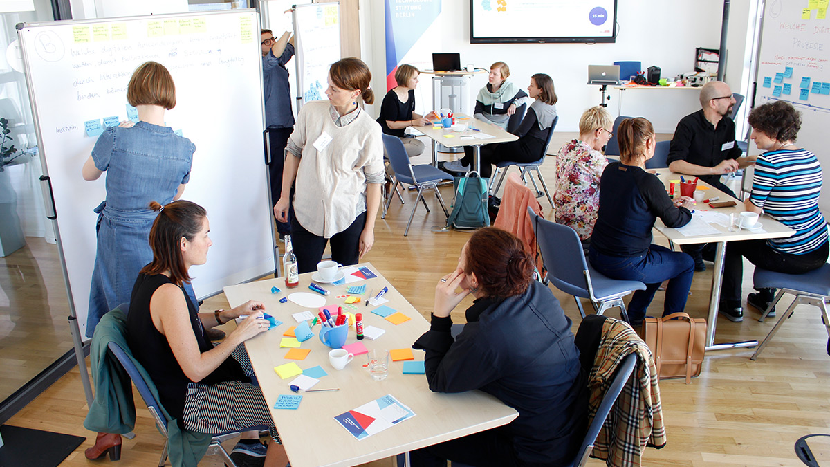 Mehrere Teilnehmer:innen eines kulturBdigital-Workshops arbeiten an Tischgruppen und Stellwänden.