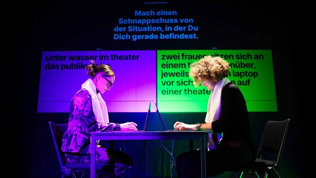 Zwei Frauen sitzen auf einer Bühne, jeweils in die Tastatur eines Laptops tippend. Hinter ihnen ist eine Aufgabe eingeblendet sowie die jeweiligen Lösungen, an denen die Frauen arbeiten.