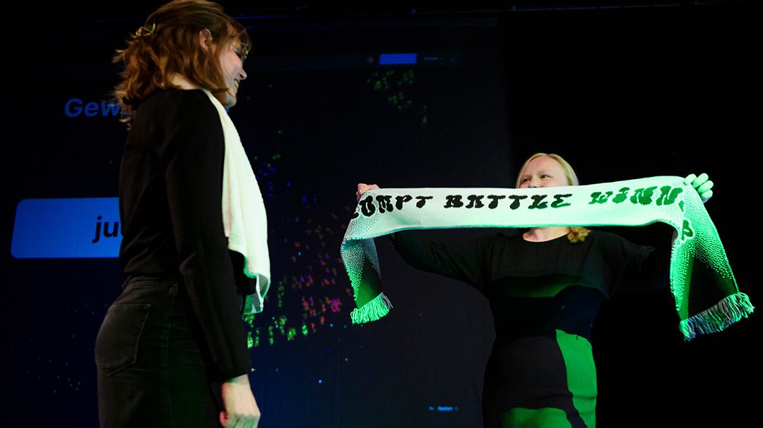 Zu sehen sind zwei junge Frauen. Die rechte Frau hält einen Schal mit der Aufschrift "Prompt Battle Winner" hoch und überreicht ihn der linken Frau.