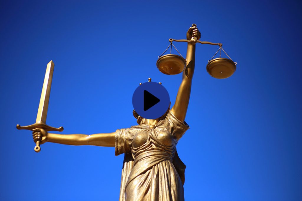 Eine Bronzeskulptur der Gerechtigkeitsgöttin Justititia vor blauem Himmel, mit verbundenen Augen einem Schwert in der einen und der Waage in der anderen Hand. In der Mitte des Bildes signalisiert ein Play-Button, dass es sich um einen Video-Beitrag handelt.