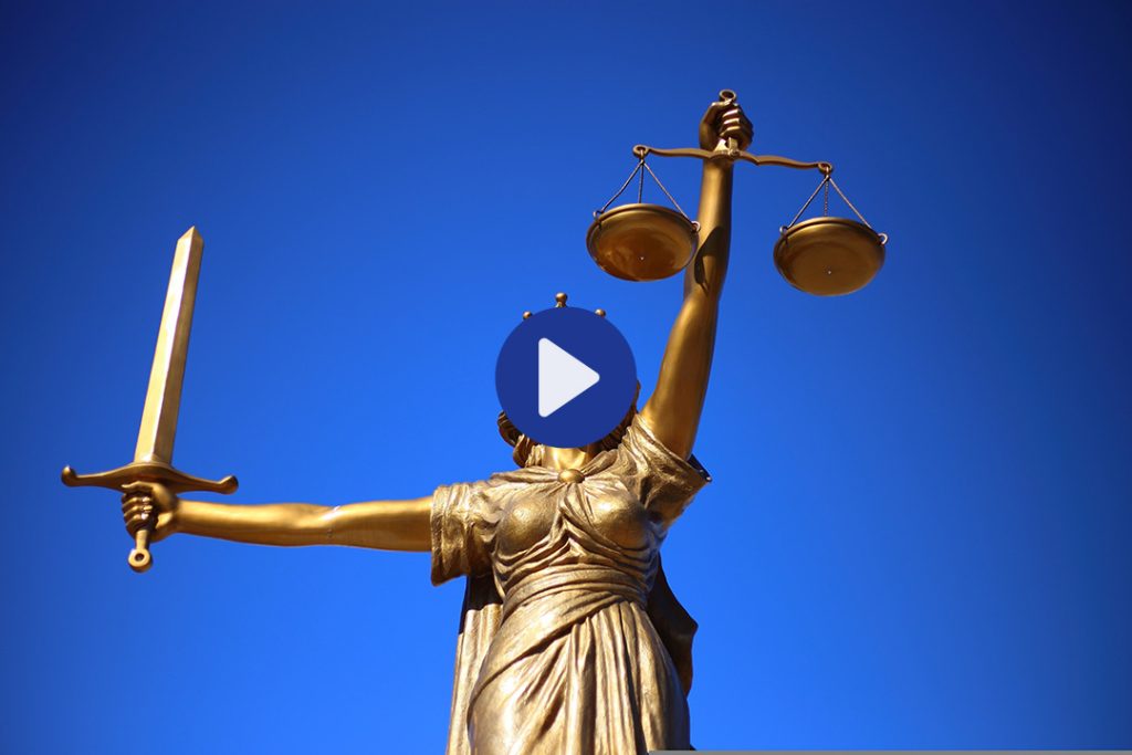 Eine Bronzeskulptur der Gerechtigkeitsgöttin Justititia vor blauem Himmel, mit verbundenen Augen einem Schwert in der einen und der Waage in der anderen Hand. In der Mitte des Bildes signalisiert ein Play-Button, dass es sich um einen Video-Beitrag handelt.