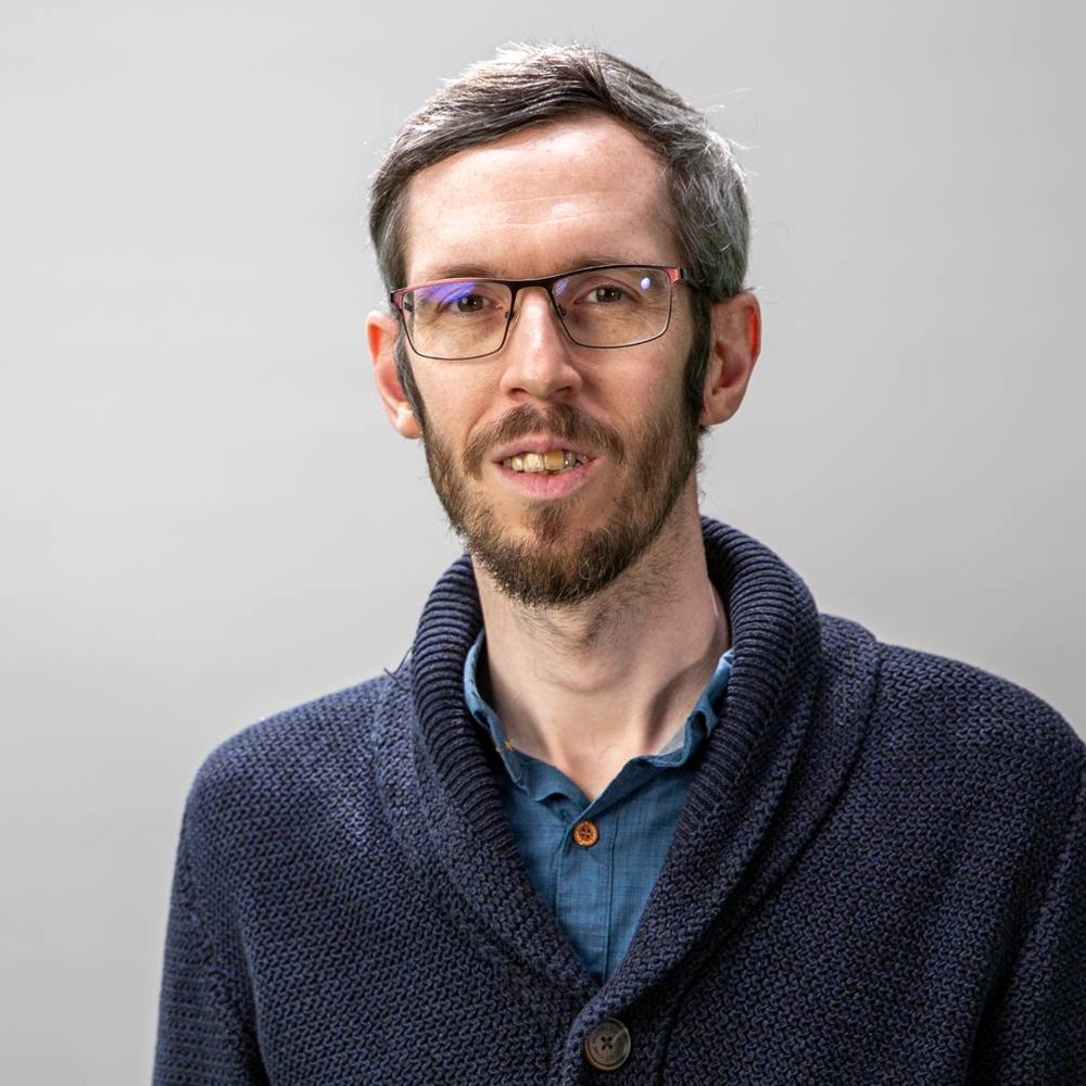 Porträt von Christoph Wetzel mit Brille und Bart vor grauem Hintergrund, in dunkelblauer Strickjacke