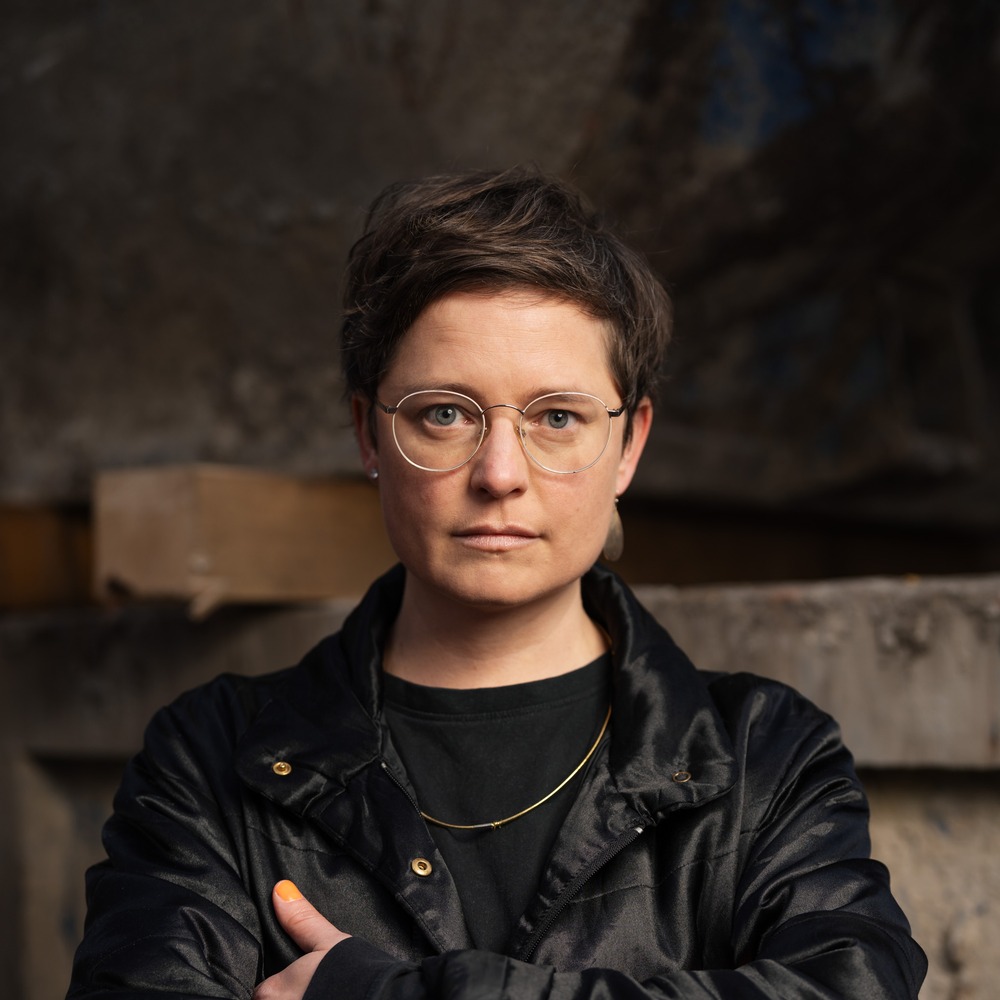 Porträt von Katharina Haverich vor dunklem Hintergrund, in schwarzer Jacke mit verschränkten Armen.